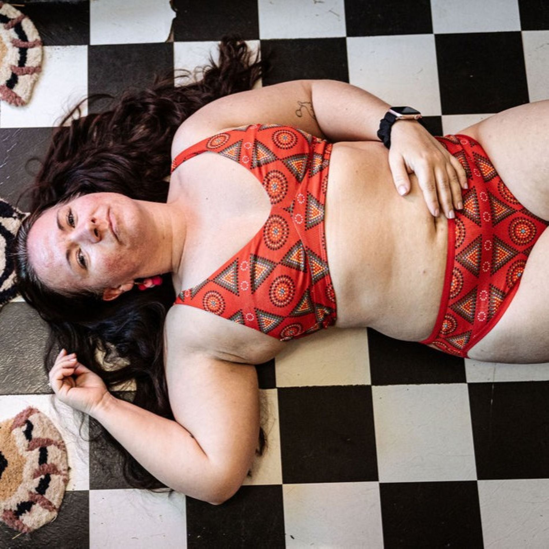 A photo of a model wearing the YOU Underwear red mara bralette and mid-rise bikini knickers. She is laying on a black and white checked floor with her left hand on her stomach and her right up by her head.