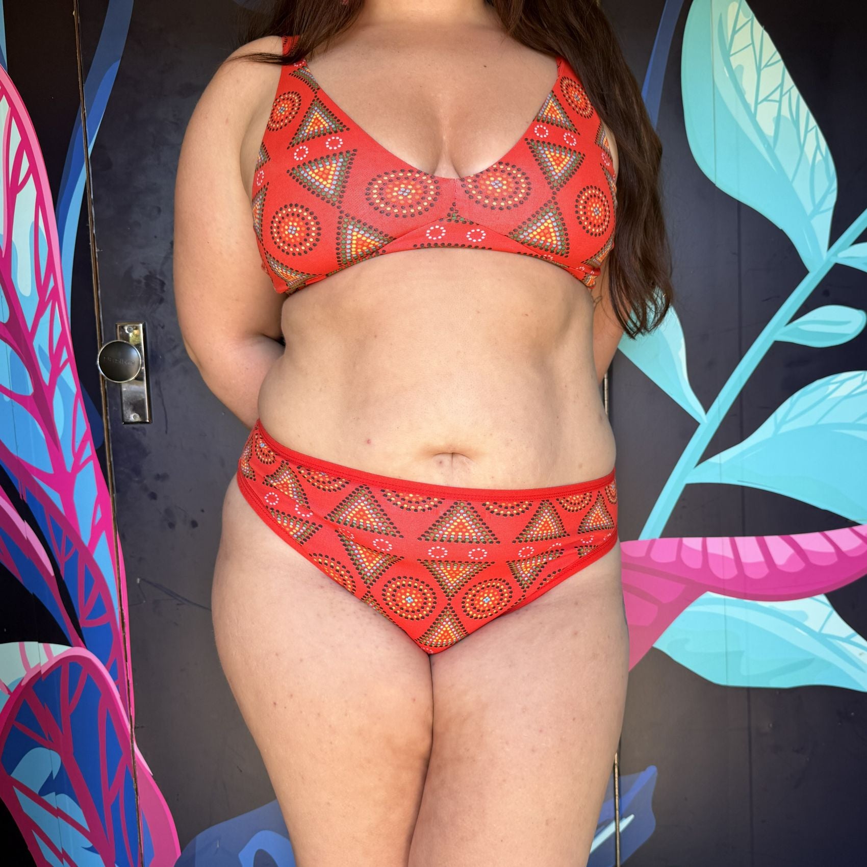 A photo of a model wearing the YOU Underwear red mara bralette and matching mid-rise knickers. The photo is zoomed in so we can see from the model's shoulders to their lower thigh. They are facing the camera, with their hands behind their back. There is a black wall with bright blue and pink leaves on as the background.