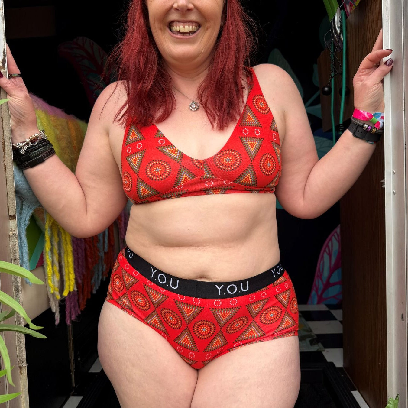 A close up photo of a model wearing the YOU Underwear bralette and boyshorts in red mara. We can see from the model's nose to her upper thigh, she is facing the camera, smiling, and has her hands on either side of a door frame.