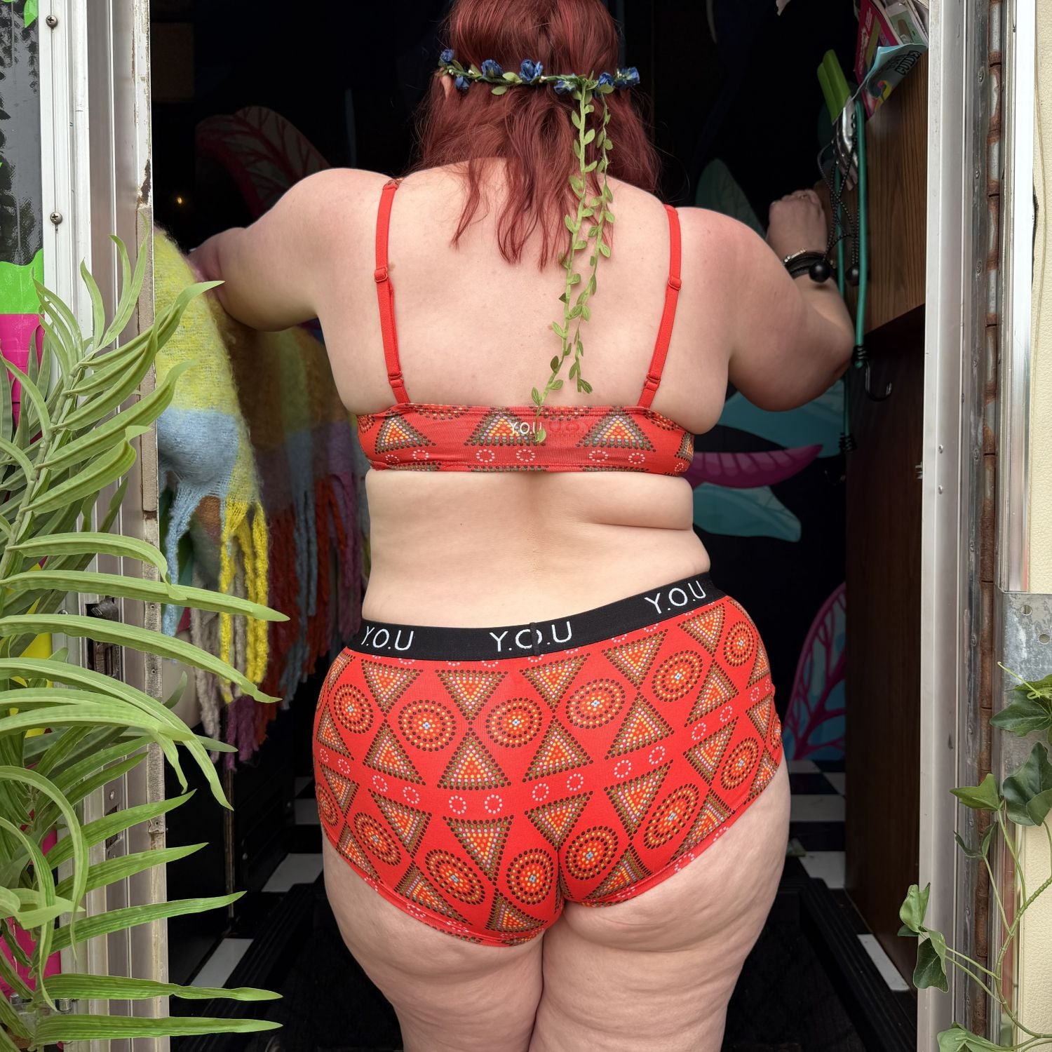 A photo of a model wearing the YOU Underwear bralette and boyshorts in red mara. She is facing away from the camera, stood in a doorways with her hands reached out in front of her. There is some green foliage in the left hand side of the photo.