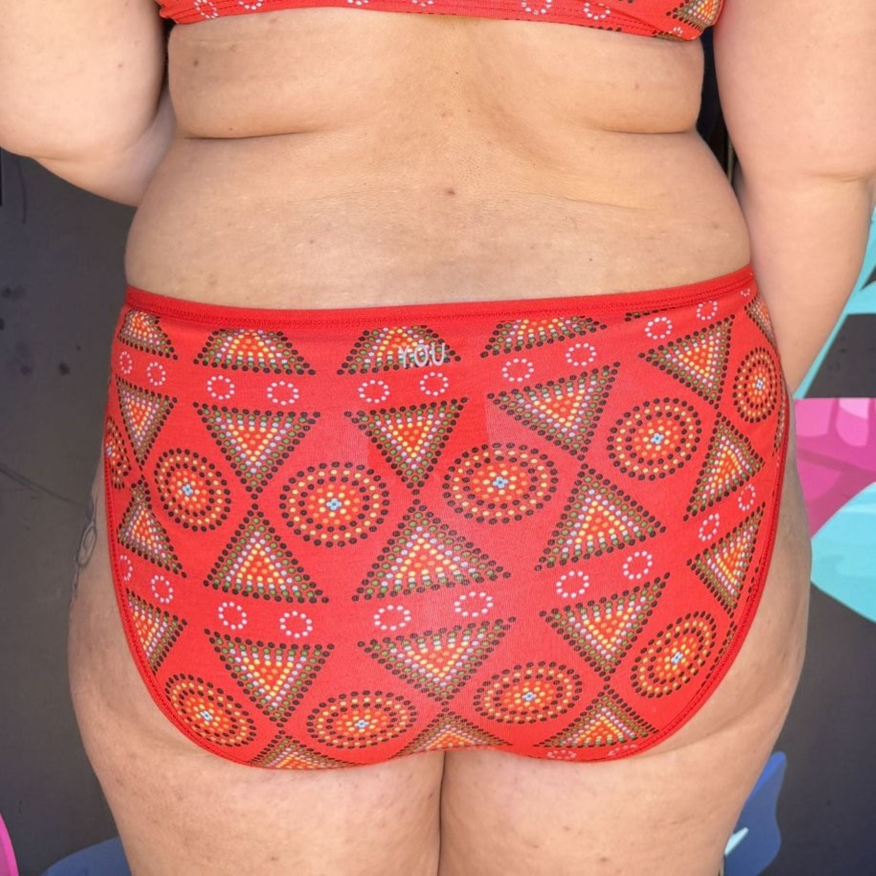 A close up photo of a model wearing the YOU Underwear mid-rise bikini knickers in red mara. She is facing away from the camera and the photo is zoomed in so we can see from the bottom of her red mara bralette to her upper thigh.