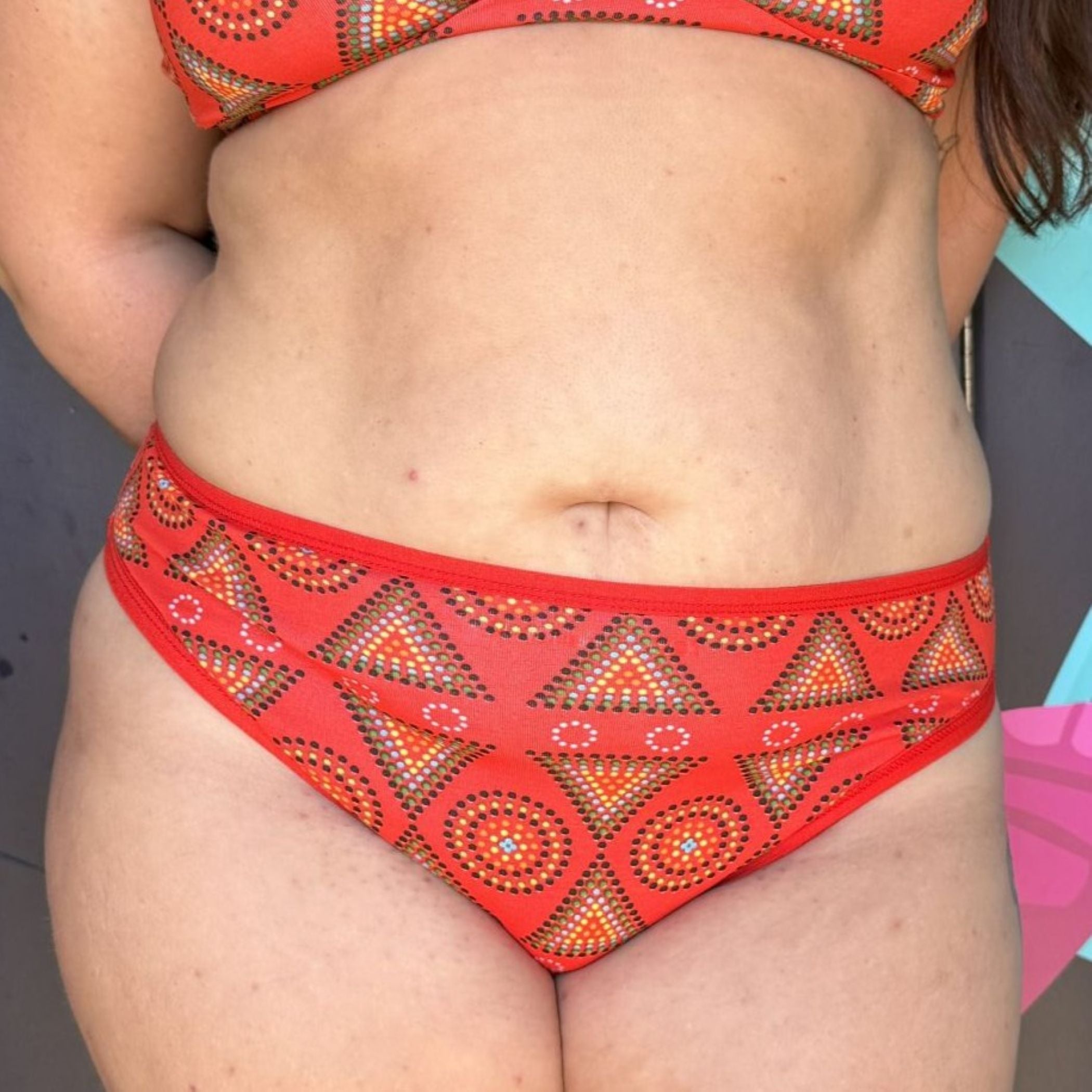 A close up photo of a model wearing the YOU Underwear mid-rise bikini knickers in red mara. She is facing the camera, with her hands behind her back and the photo is zoomed in so we can see from the bottom of her red mara bralette to her upper thigh.