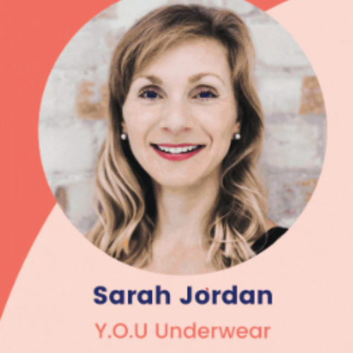An image of Sarah Jordan, the founder of YOU Underwear. She has blonde hair and is wearing red lipstick with pearl earrings. The background of the image is light pink and bright coral.