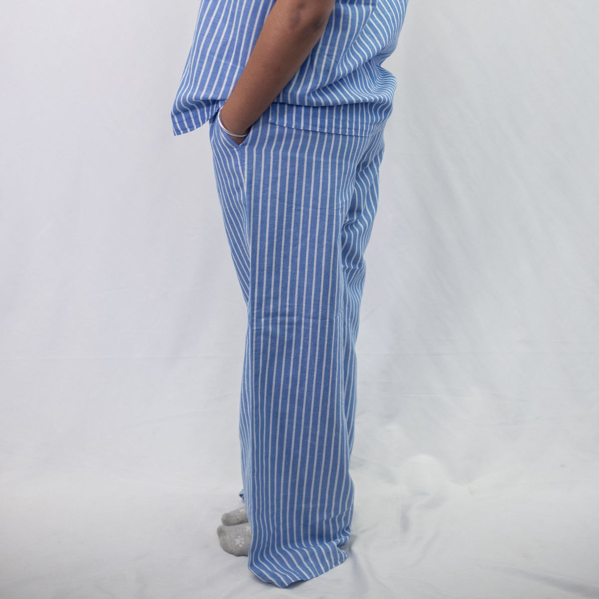 A photo of a model wearing the YOU Underwear woven pyjama bottoms in the classic blue and white stripe. The photo is zoomed in so we can see from the model's stomach to the floor. They are standing side onto the camera with their hands in their pockets and there is a white background behind them.