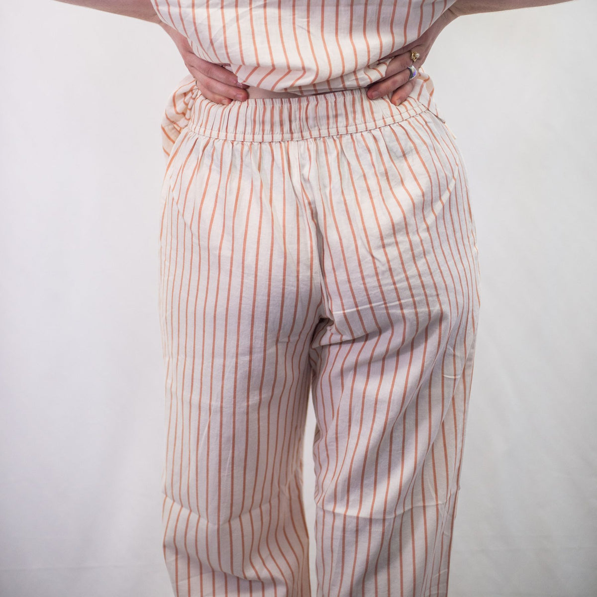 A photo of a model wearing the YOU Underwear woven pyjama bottoms in soft cream and coral. The photo is zoomed in so we can see from the model's stomach to their shins. They are facing away from the camera and holding up their shirt so we can clearly see the trouser waistband. There is a white background.