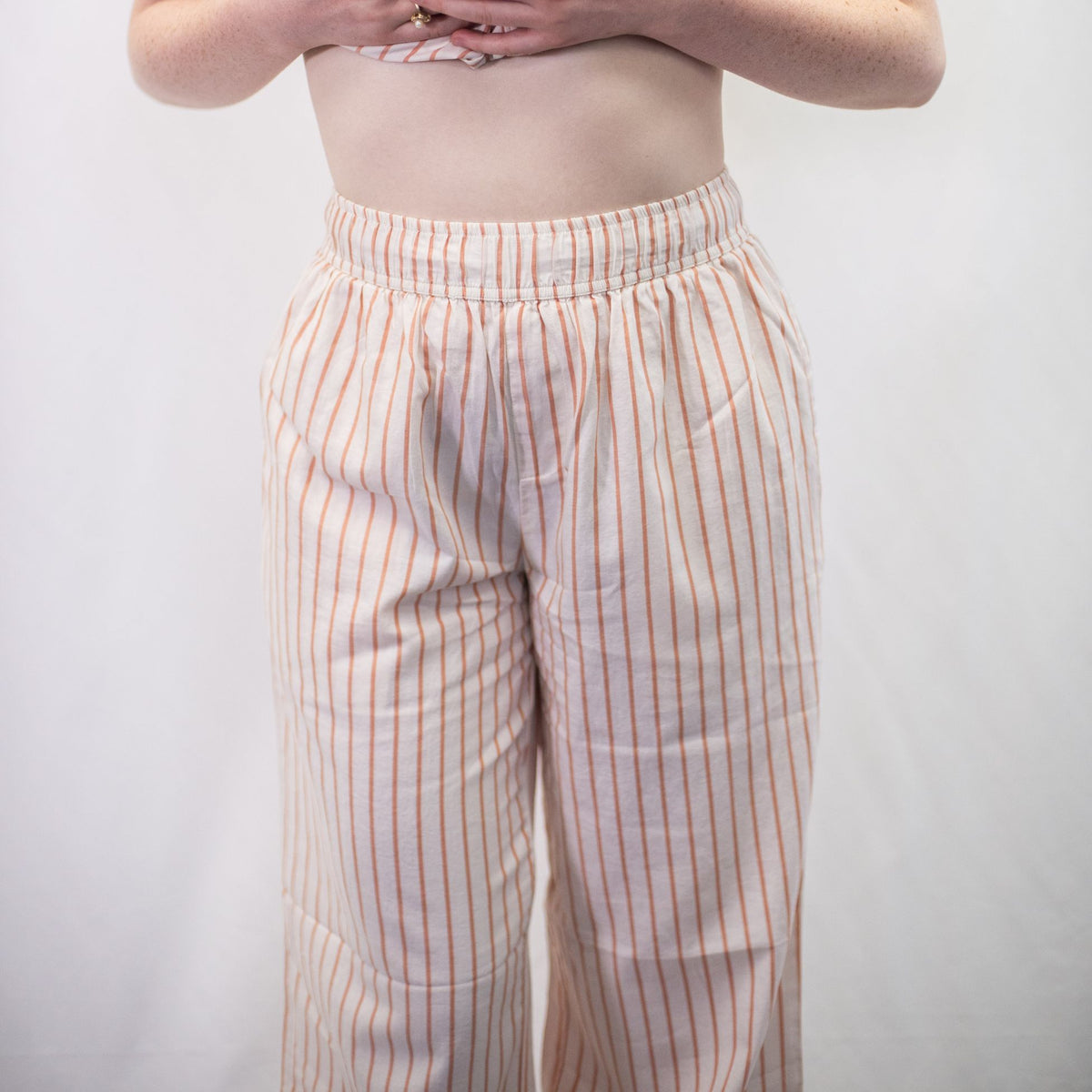 A photo of a model wearing the YOU Underwear woven pyjama bottoms in soft cream and coral. The photo is zoomed in so we can see from the model's stomach to their shins. They are facing the camera and holding up their shirt so we can clearly see the trouser waistband. There is a white background.