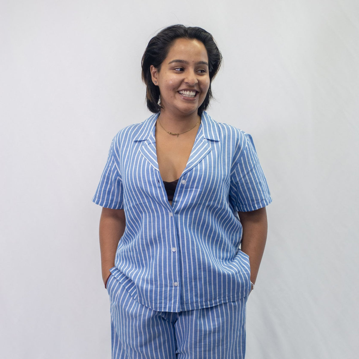 A photo of a model wearing the YOU Underwear woven pyjama shirt in the classic blue and white stripe. She is standing and facing the camera, looking off the left and smiling. There is a white background behind her.