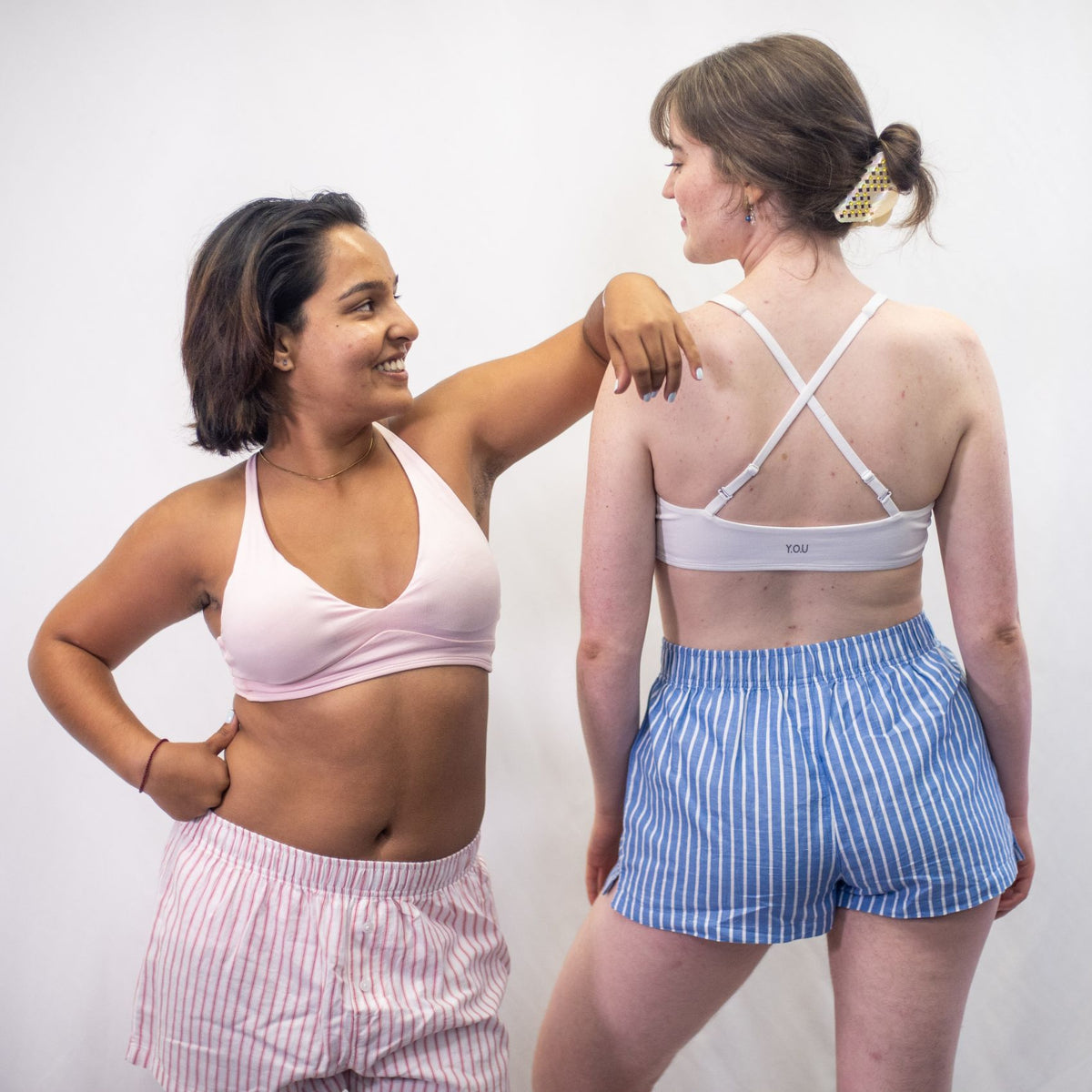 A photo of two women wearing YOU Underwear bralettes and the woven pyjama shorts. The model on the left is wearing the light pink bralette and the wpink and white striped woven shorts and is facing the camera, leaning on the other model with her left hand. The model on the right is wearing the white bralette and the blue and white stripe pyjama shorts. She is facing away from the camera with her hands at her sides. There is a white background.