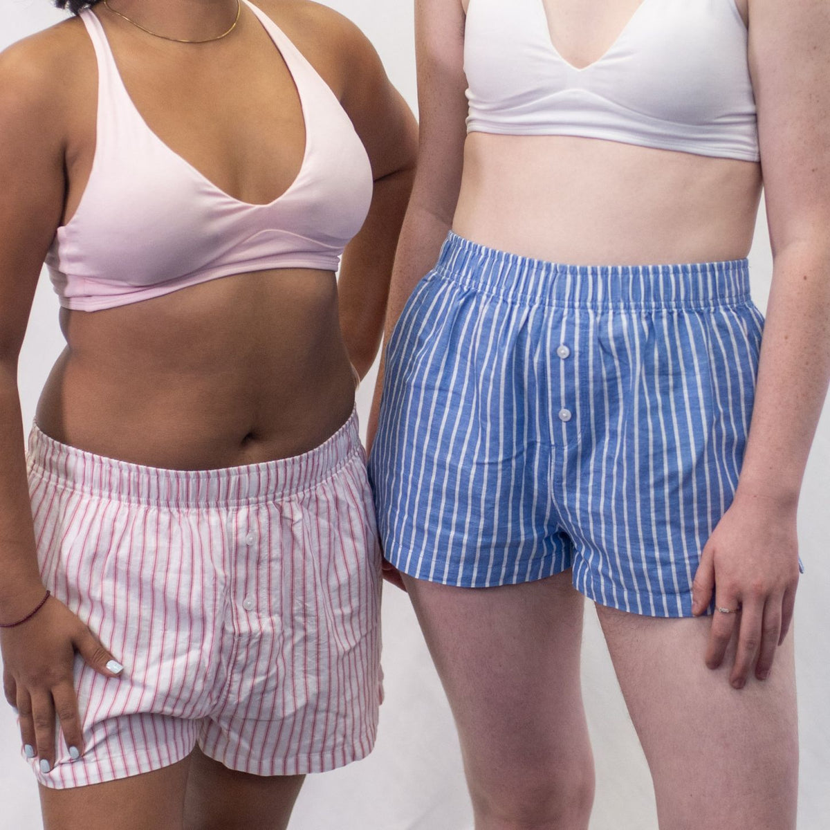 A close up photo of two models who are wearing the YOU Underwear woven pyjama shorts. The woman on the left is wearing a pink bralette and white and pink stripe shorts and the woman on the right is wearing a white bralette and blue and white stripe pyjama shorts. There is a white background behind them and they are both standing facing the camera.