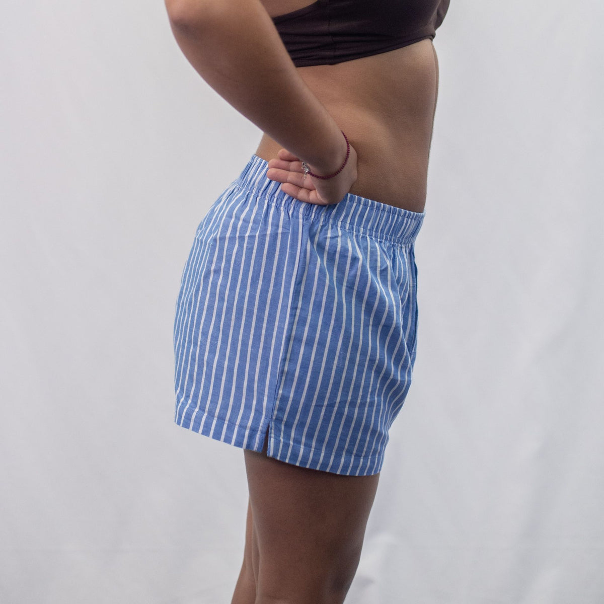 A photo of a model wearing the YOU Underwear woven pyjama shorts in the classic blue and white stripe. The photo is zoomed in so we can see from the model's mid-stomach to her knees. She is standing side on to the camera and has her hands on her hips. There is a white background.