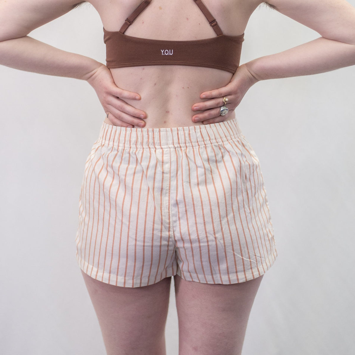 A photo of a model wearing the YOU Underwear woven pyjama shorts in the soft cream and coral stripes. The photo is zoomed in so we can see from the model's upper-back to their upper-thigh. She is facing away from the camera and has her hands on her hips. There is a white background.