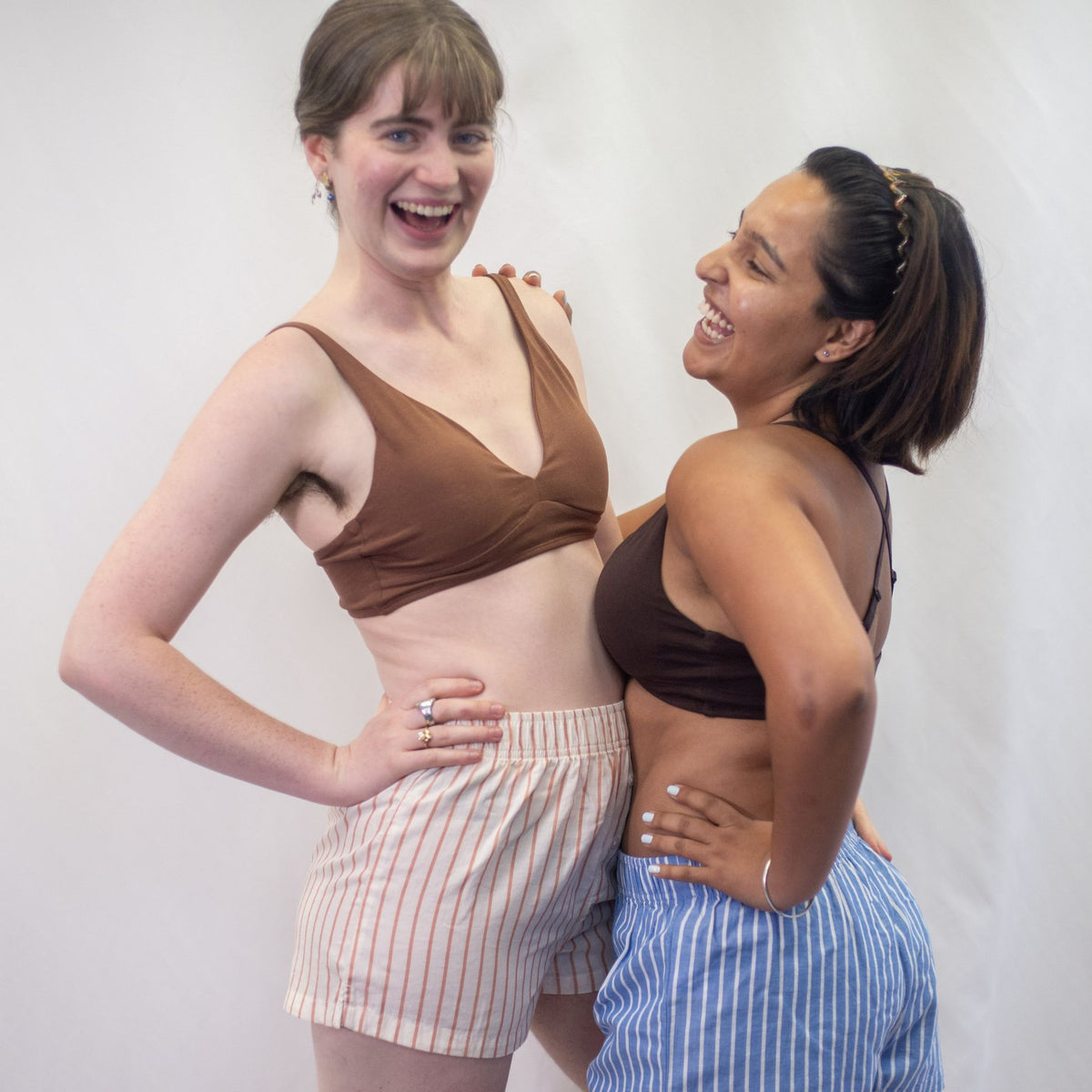 A photo of two models wearing YOU Underwear bralettes and woven pyjama shorts. They are standing closely together embracing each other with one arm. The model on the left is wearing a chestnut bralette and cream and coral striped pyjama shorts. The model on the right is wearing a cocoa bralette and blue and white striped pyjama shorts. They appear to be laughing and each have one hand on their hips. There is a white background.