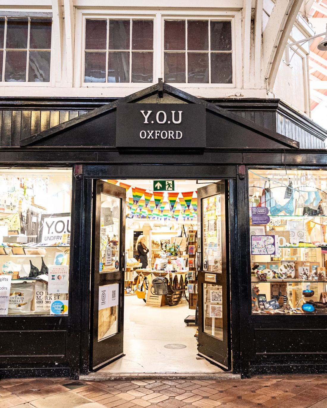 A photo of the outside of the YOU Oxford shop. The building's structure is black and there are double doors which are open, inviting people into the shop. The windows are big and have colourful displays in them. We can see that there is pride bunting hang up in the shop.
