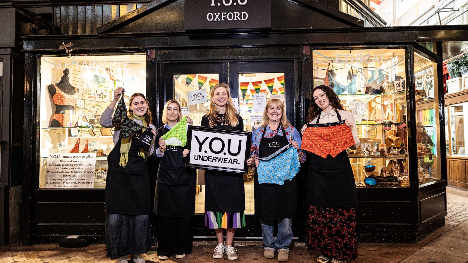 A photo of the YOU Oxford shop team standing outside the shop, all holding underwear. There are five women and the woman in the middle is holding a big black and white sign which reads, 'YOU Underwear'. They are stood in a line, facing the camera and smiling. The YOU Oxford shop is behind them, with black infrastructure and big windows with colourful displays in them.