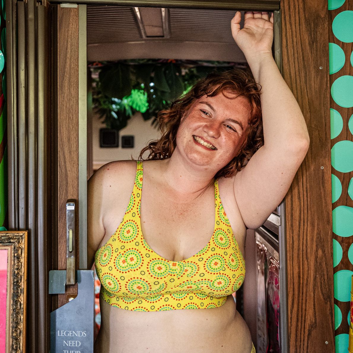 A photo of a model wearing the YOU Underwear non wired bra in yellow mara. She is standing facing the camera, leaning against a doorway with her left hand on the door frame. The photo is zoomed in so we can see to the model's waist.