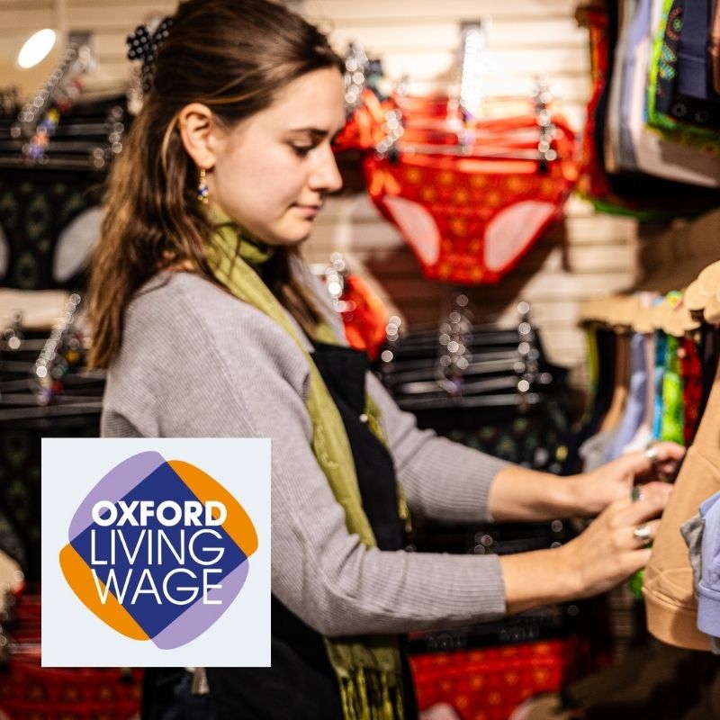 a woman wearing a black YOU apron sorts and tidies the display in the YOU Oxford shop with an Oxford Living Wage logo in the corner.