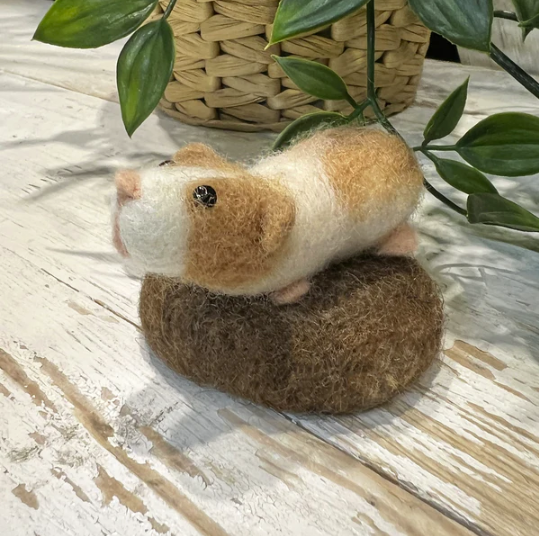 needle felted guinea pig on felted block with a plant in a pot behind it