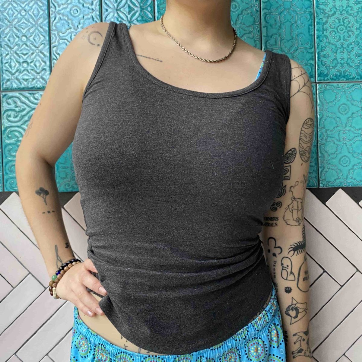 A close up photo of a model wearing the YOU Underwear vest in charcoal grey. They are facing the camera, with their left hand on her hip. They are stood in front of a white, pink and blue mosaic wall.