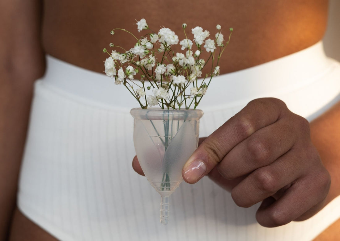 A woman wearing a lilac bra and sage underwear lays on a bed holding a reusable menstrual cup