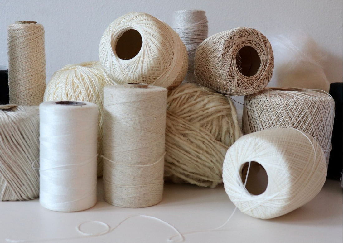 11 white, cream and ivory coloured spools of cotton and wool are jumbled together on a white table