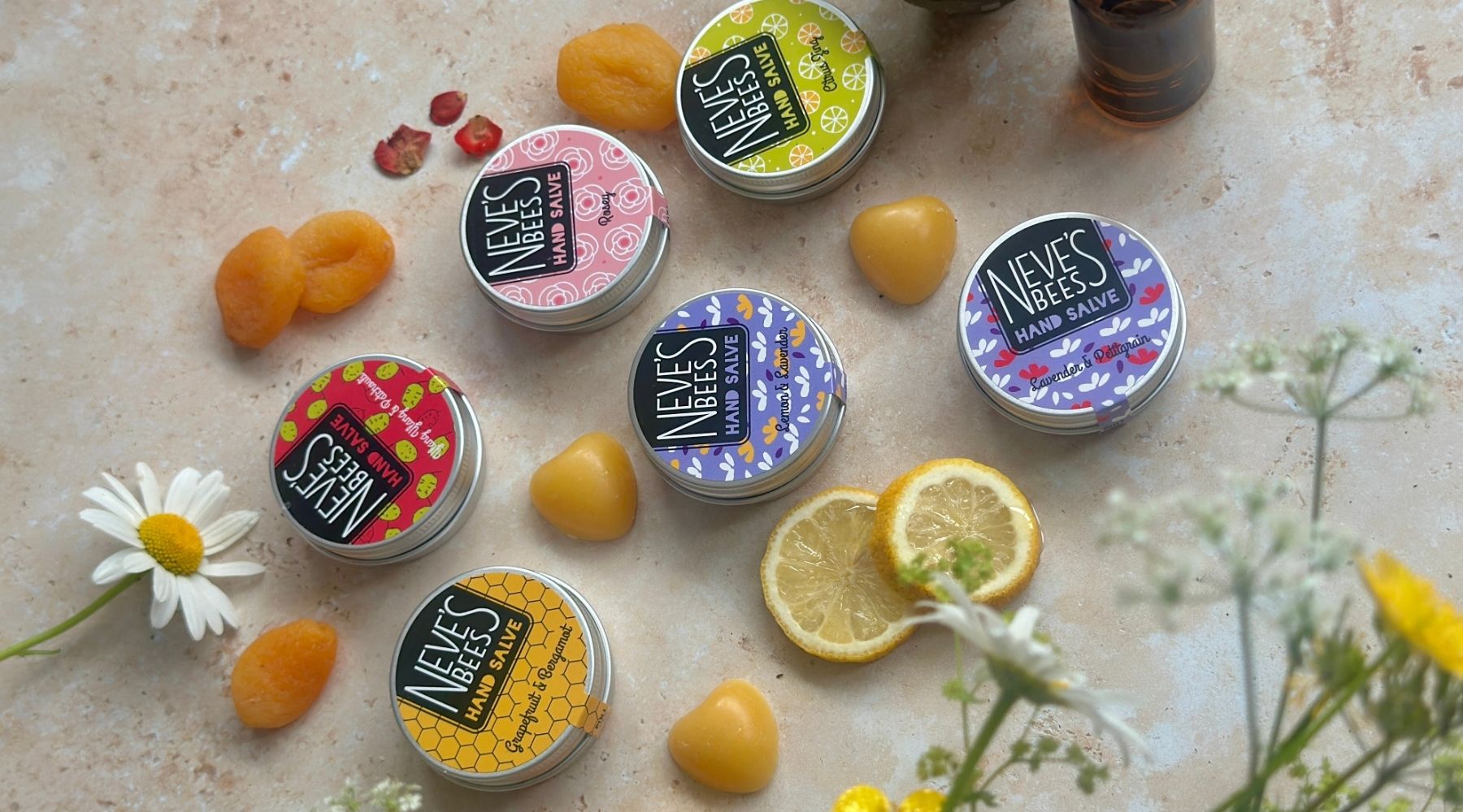 containers of Neve's Bees hand salve arranged on a stone coloured background with fruits laid around.