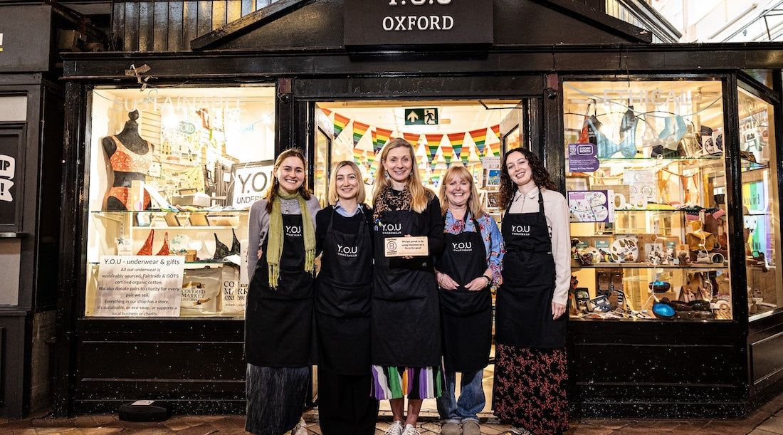 fine women wearing YOU aprons stand outside the Oxford YOU shop. The woman in the middle is holding a wooden B Corp sign.