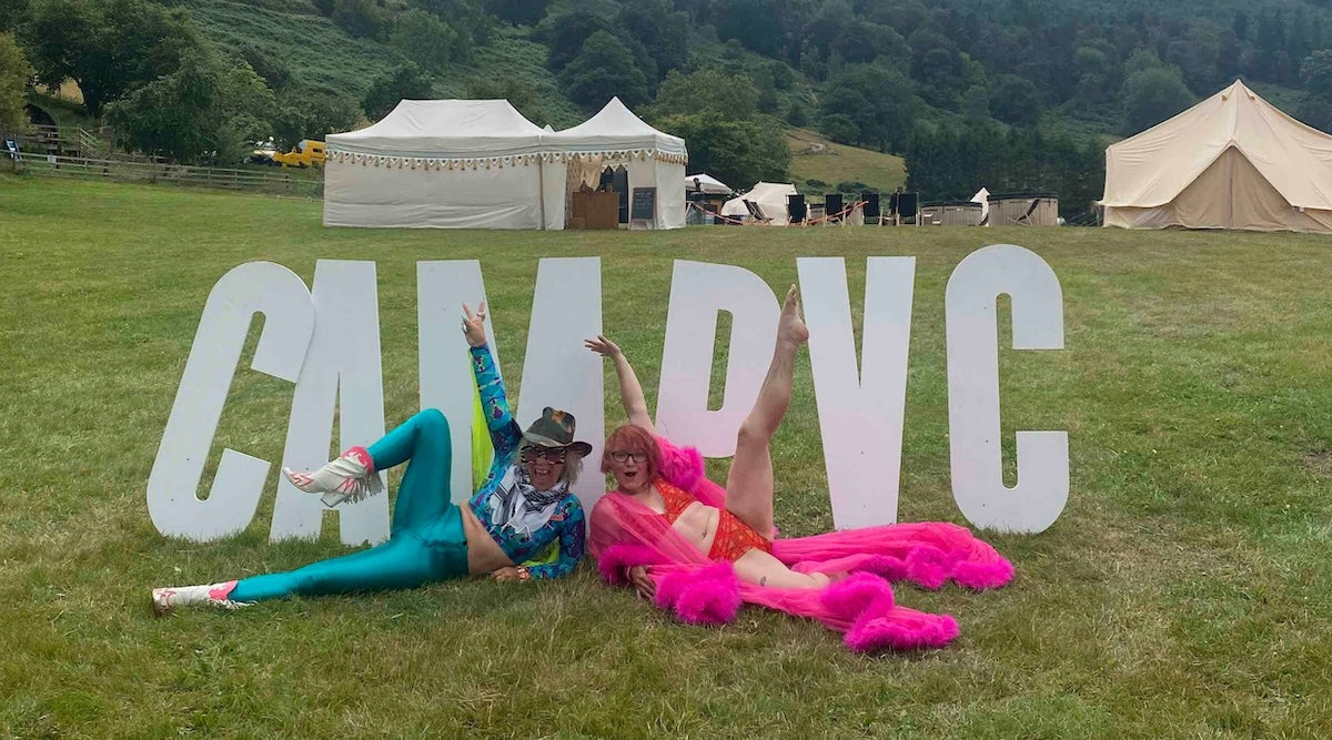 a big white Camp VC letters sign in a field with two women lying down to pose in front of it. One woman is wearing turquoise blue trousers and top and the other wears YOU red mara matching set with a pink robe.