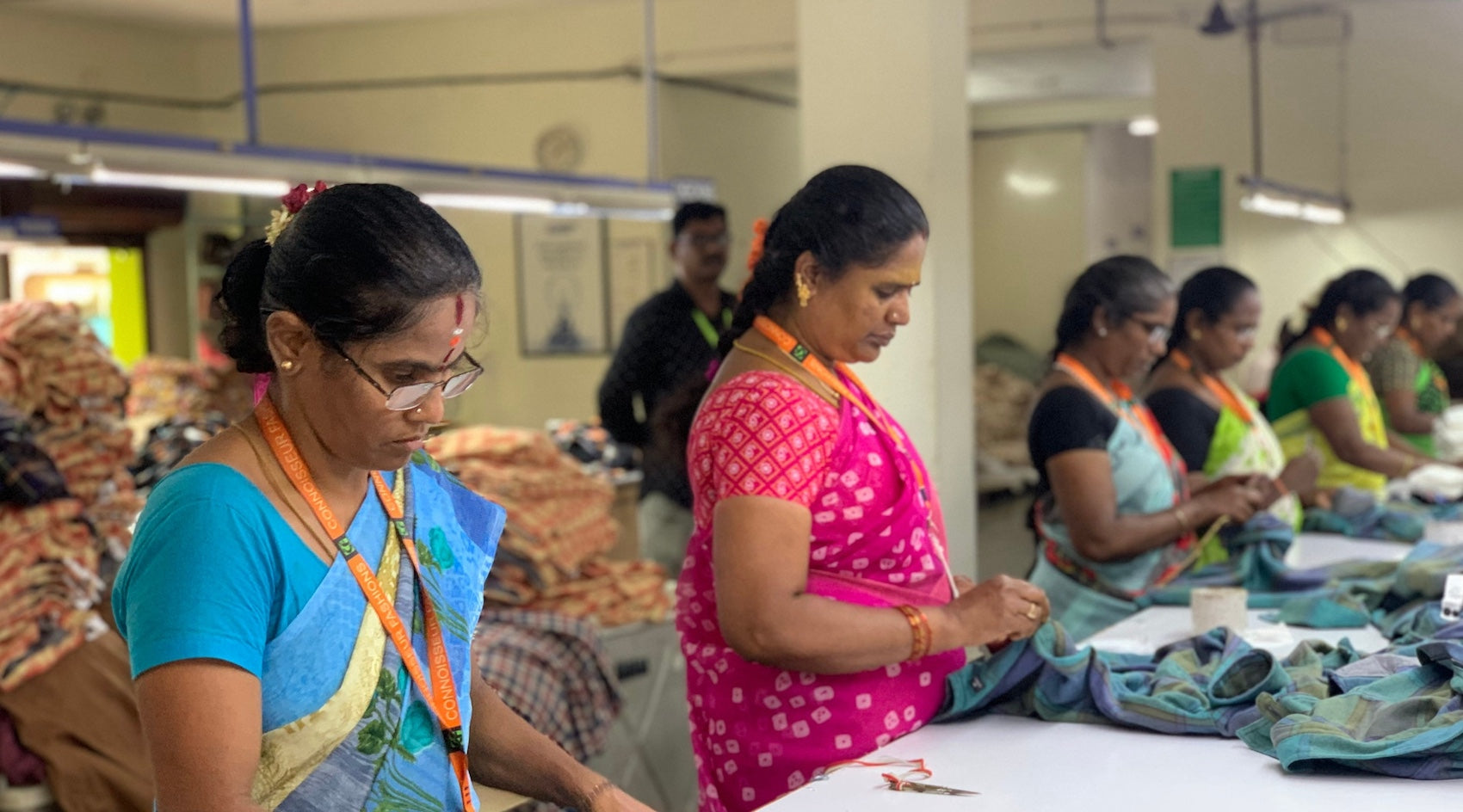 two women wearing saris stand at a work bench inside a factory inspecting clothing