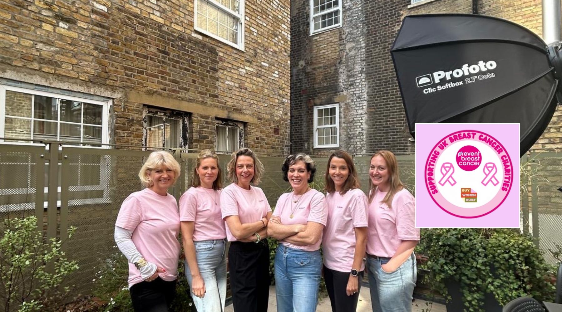 six women wearing pink t shirts stand in a building courtyard to pose for a photo. The Buy Women Built with Prevent Breast Cancer logo is in the corner
