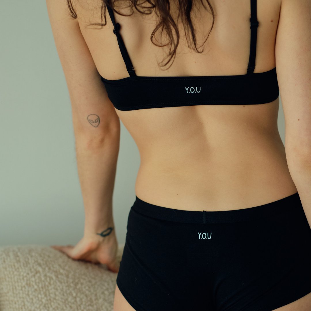 A brunette faces away from the camera wearing a black boy shorts and bralette matching set