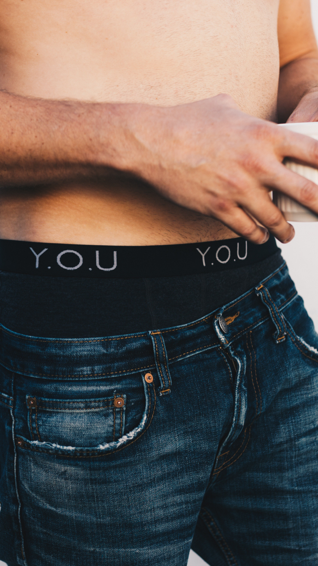 A man holding a white mug wearing black y.o.u boxers and dark blue jeans