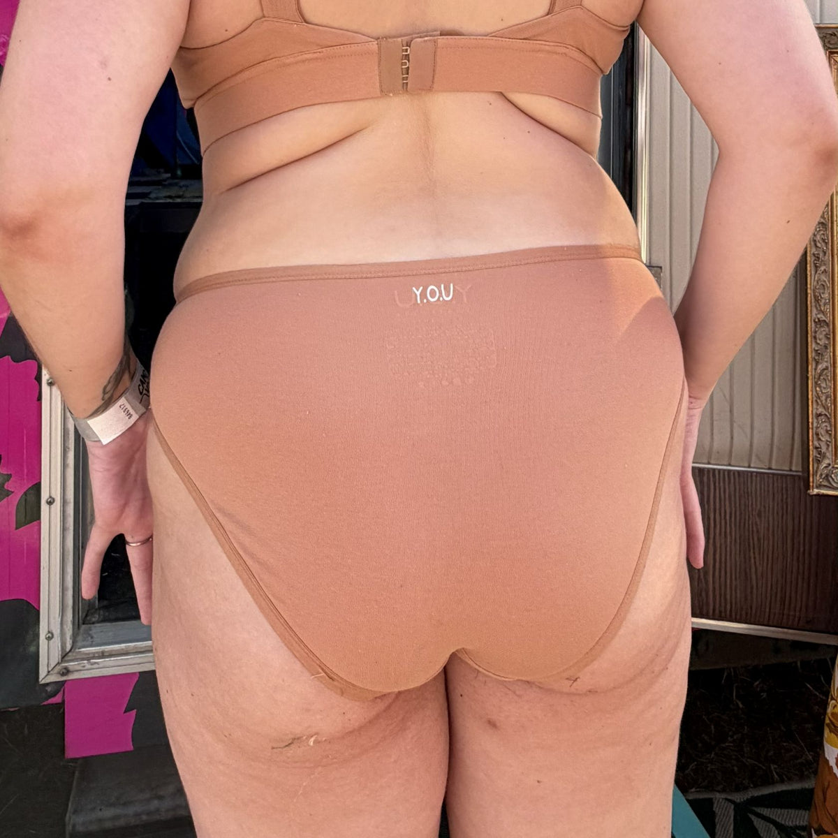 A photo of a model wearing the YOU Underwear mid rise bikini knickers in almond. They are standing facing away from the camera with their hands at their sides. The photo is cropped so we can see from the model's bust to their lower-thigh. Behind the model there is a bright pink and black leopard print bus.
