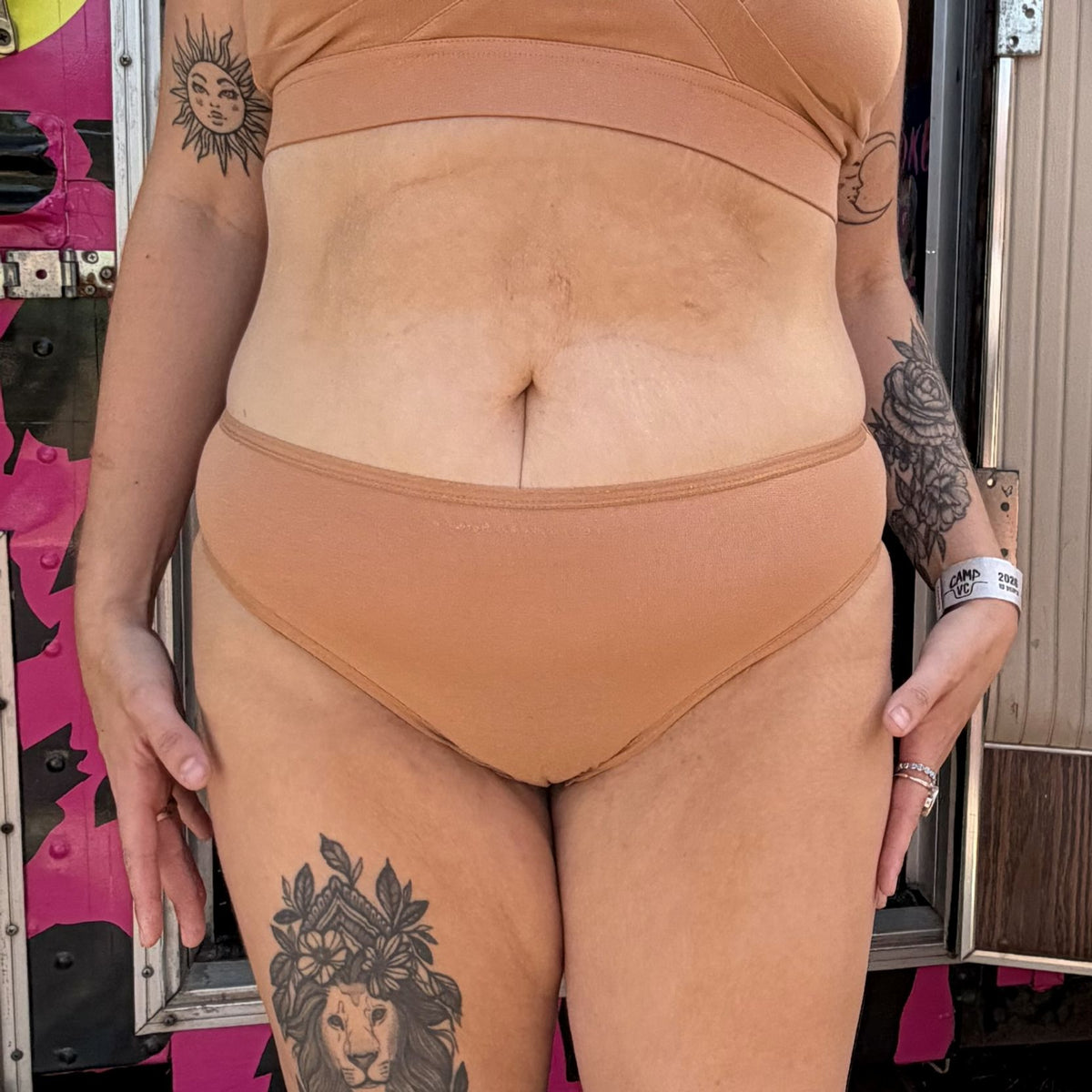 A photo of a model wearing the YOU Underwear mid rise bikini knickers in almond. They are standing facing the camera with their hands at their sides. The photo is cropped so we can see from the model's bust to their lower-thigh. Behind the model there is a bright pink and black leopard print bus.