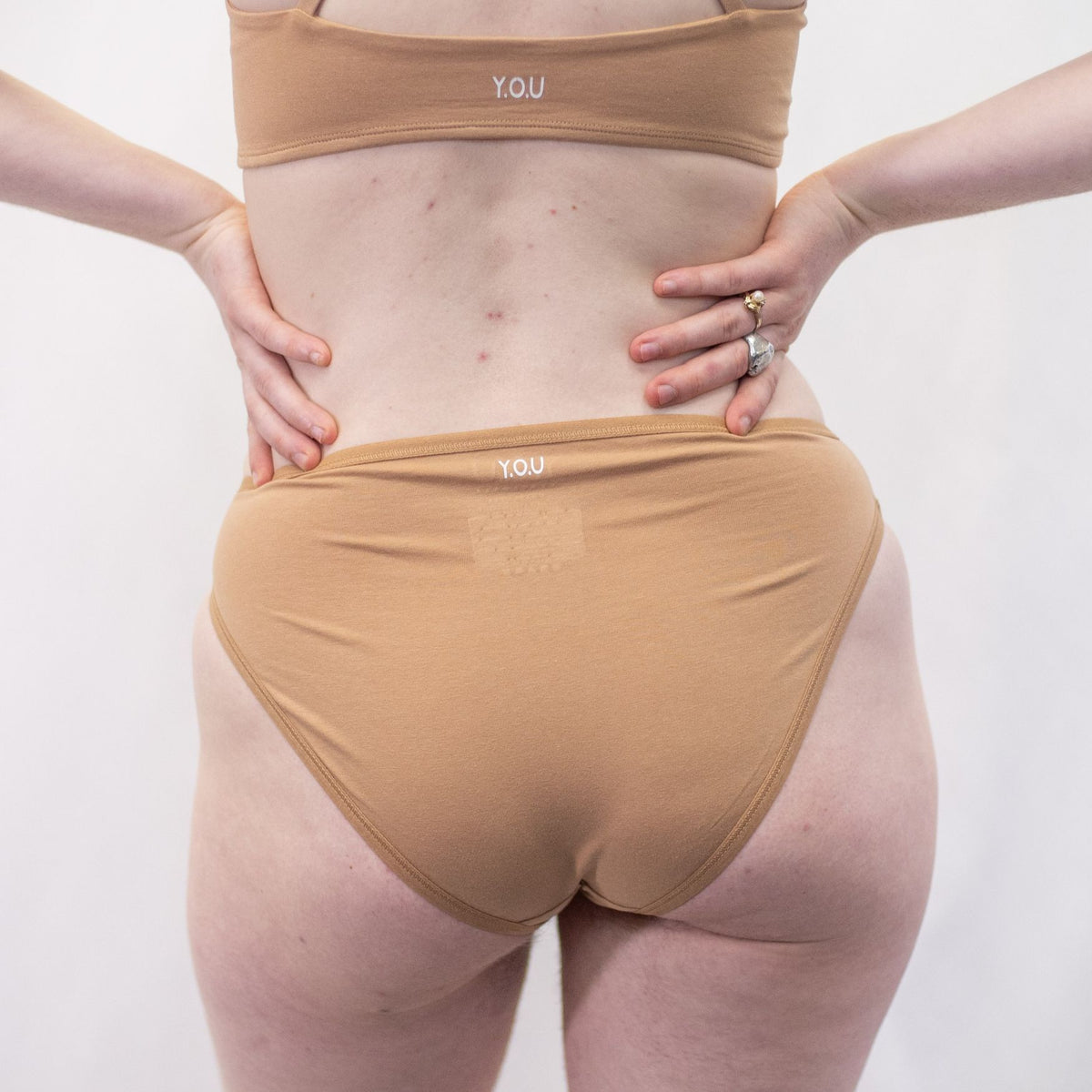 A photo of a model wearing the YOU Underwear midrise bikini knickers in almond. The model is standing facing away from the camera with their hands on their hips. The photo is zoomed in so we can see from the model's stomach to their upper thigh. The background is white.