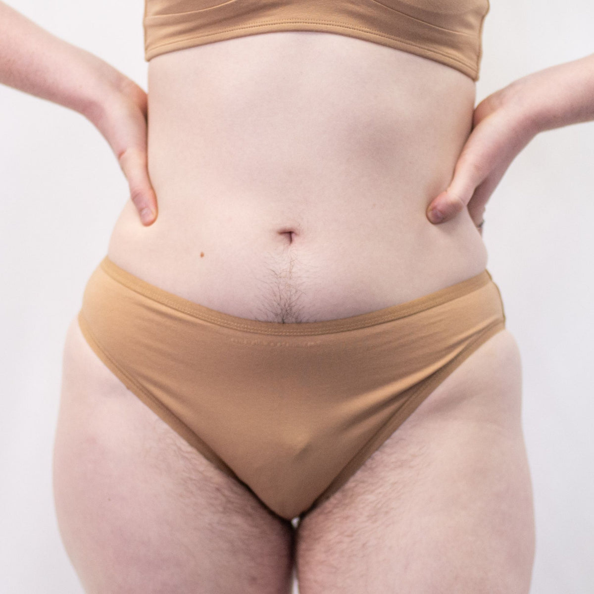 A photo of a model wearing the YOU Underwear midrise bikini knickers in almond. The model is standing facing the camera with their hands on their hips. The photo is zoomed in so we can see from the model's stomach to their upper thigh. The background is white.