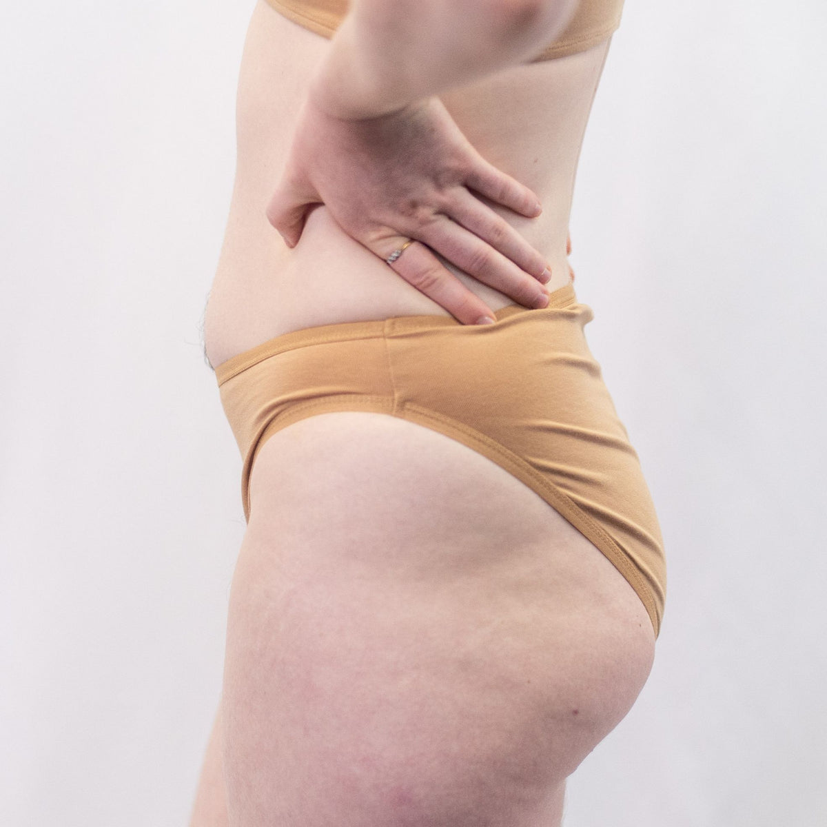 A photo of a model wearing the YOU Underwear midrise bikini knickers in almond. The model is standing side on to the camera with their hands on their hips. The photo is zoomed in so we can see from the model's stomach to their upper thigh. The background is white.