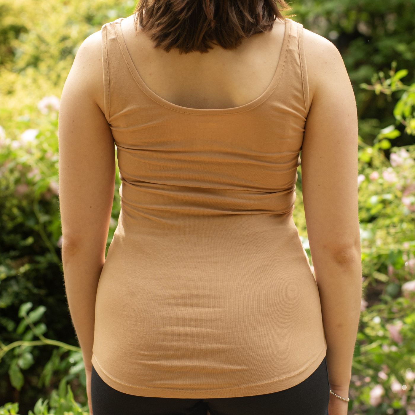 A close up photo of a model wearing the YOU Underwear almond vest. She is facing away the camera, with her hands at her sides. The photo is zoomed in so we can see from her chin to her hips. There is a green bush with pink flowers on behind her.
