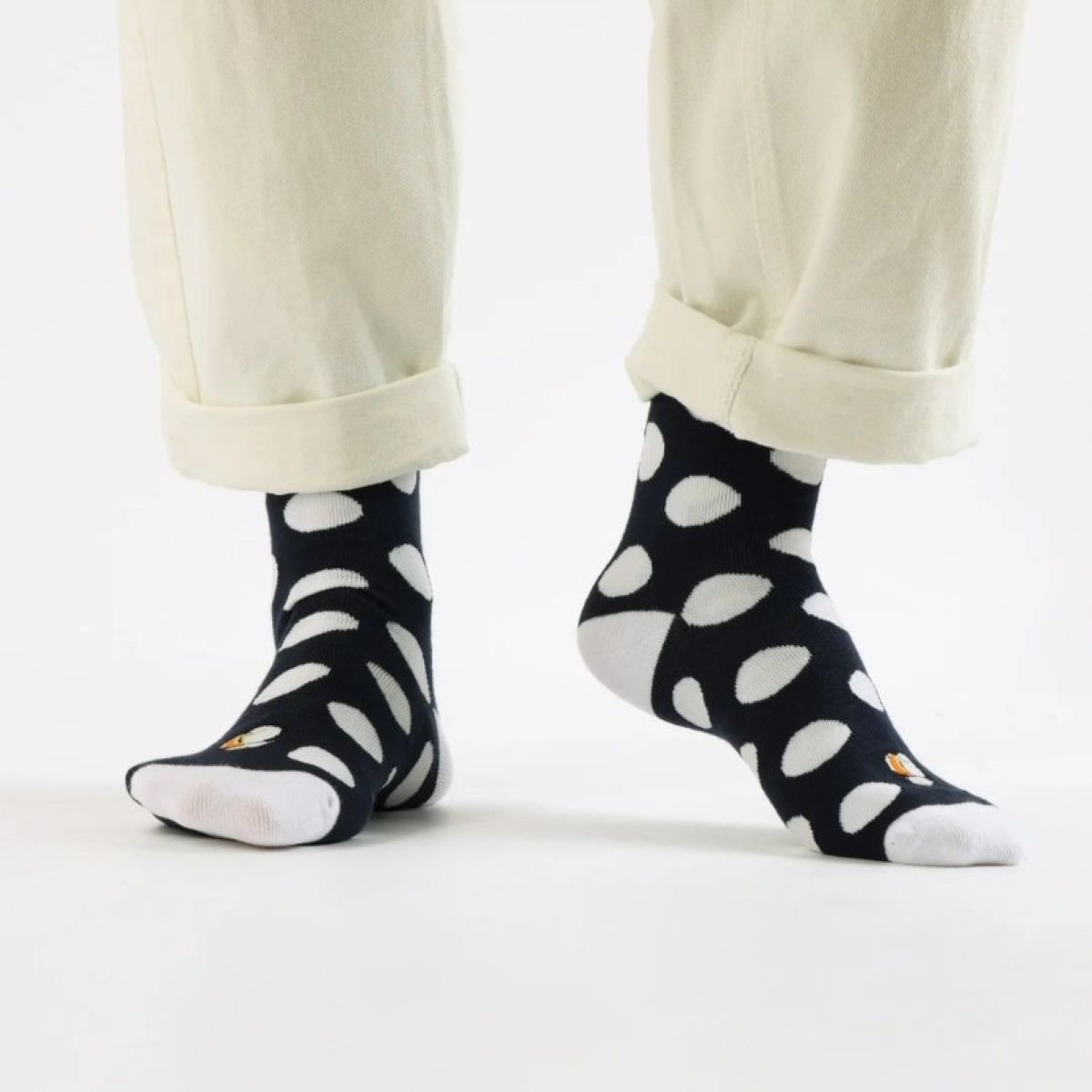 A picture of a model wearing the Bare Kind polka dot puffin socks. The picture is zoomed in so we can only see the models lower-shins and feet. They are wearing the black and white polka dot socks paired with cream jeans which are rolled up at the hem. The background is white.