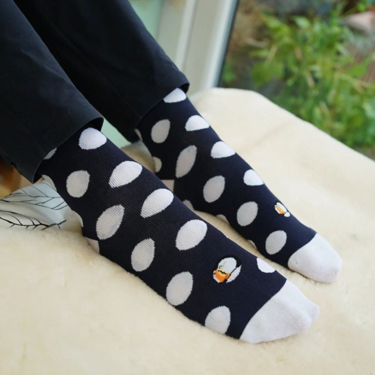 A picture of a model wearing the Bare Kind polka dot puffin socks. The picture is zoomed in so we can only see the model's lower-shins and feet. They have their feet up on a cream sofa, showing the sock's black and white polka dots along with the puffin located on the top centre of the sock. There is a garden in the background.