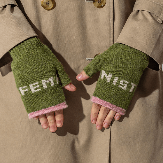 A photo of a model wearing the Black & Beech Feminist mittens in pistachio green with white writing and light pink cuffs. The model is also wearing a beige trench coat with tortoise shell buttons.