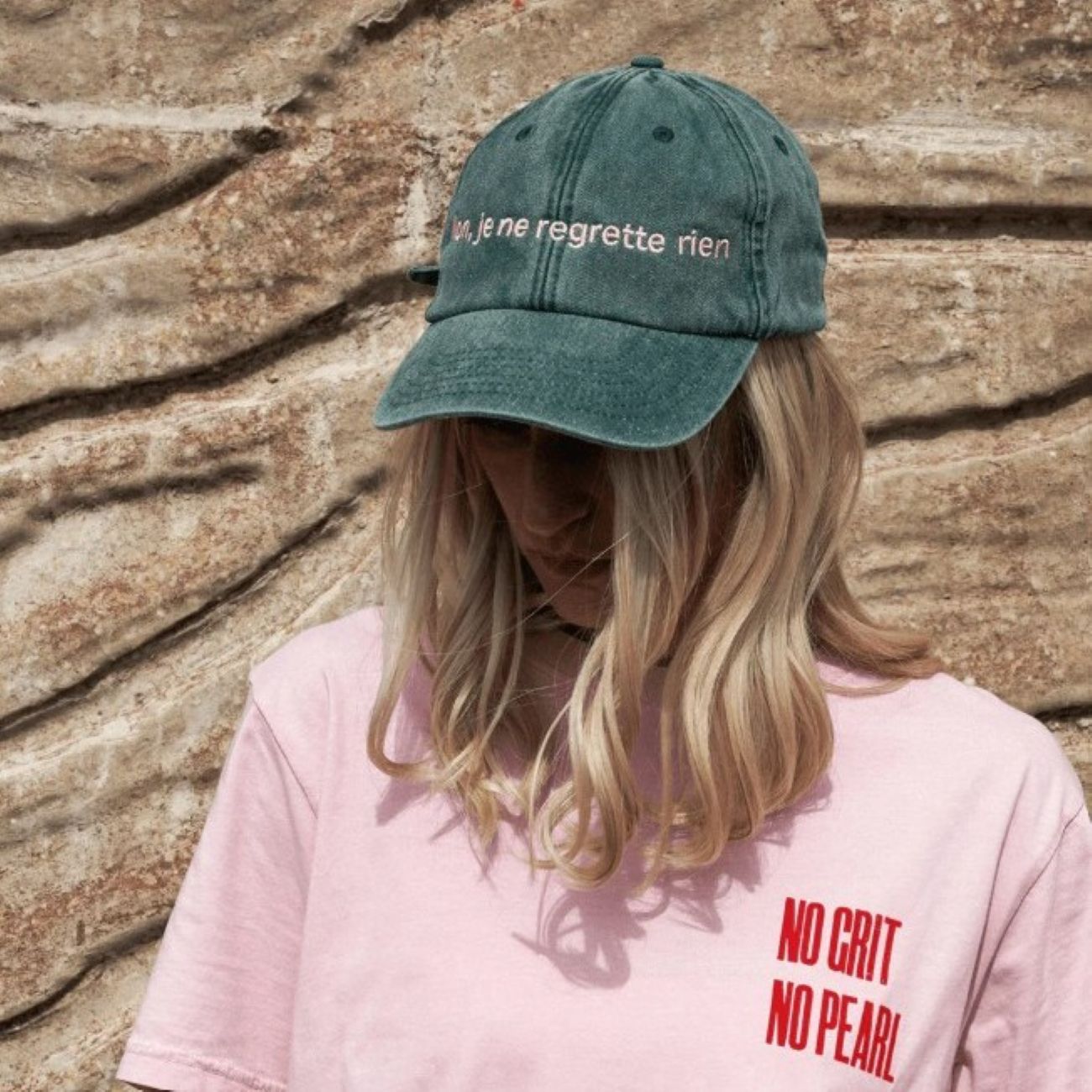 A woman wearing the Black & Beech Regrette Cap in green, standing in front of a stone wall