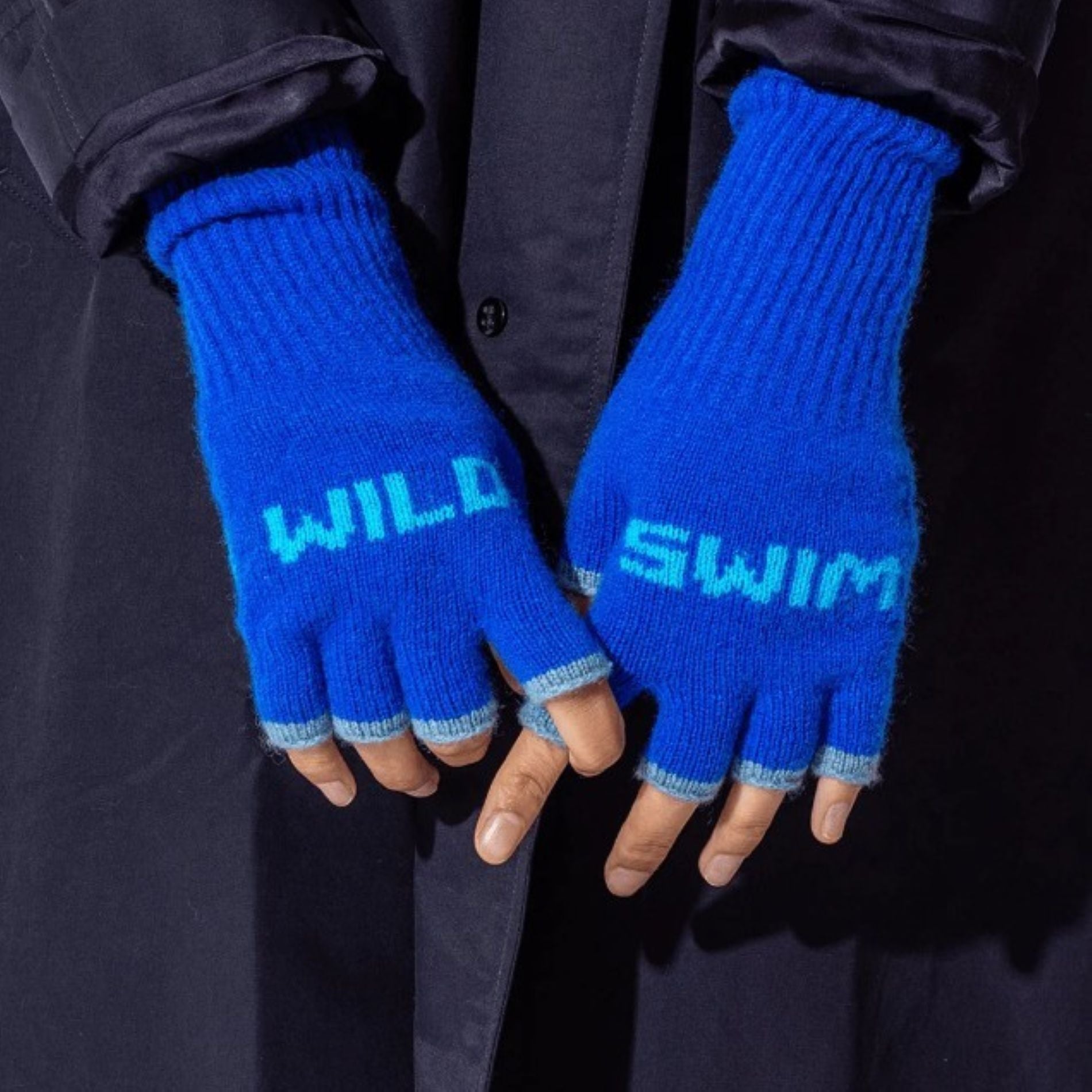 A close up photo of a model wearing the Black & Beech Wild Swim gloves in bright blue with baby blue writing and cuffs. The model is wearing a dark blue, long trench coat.