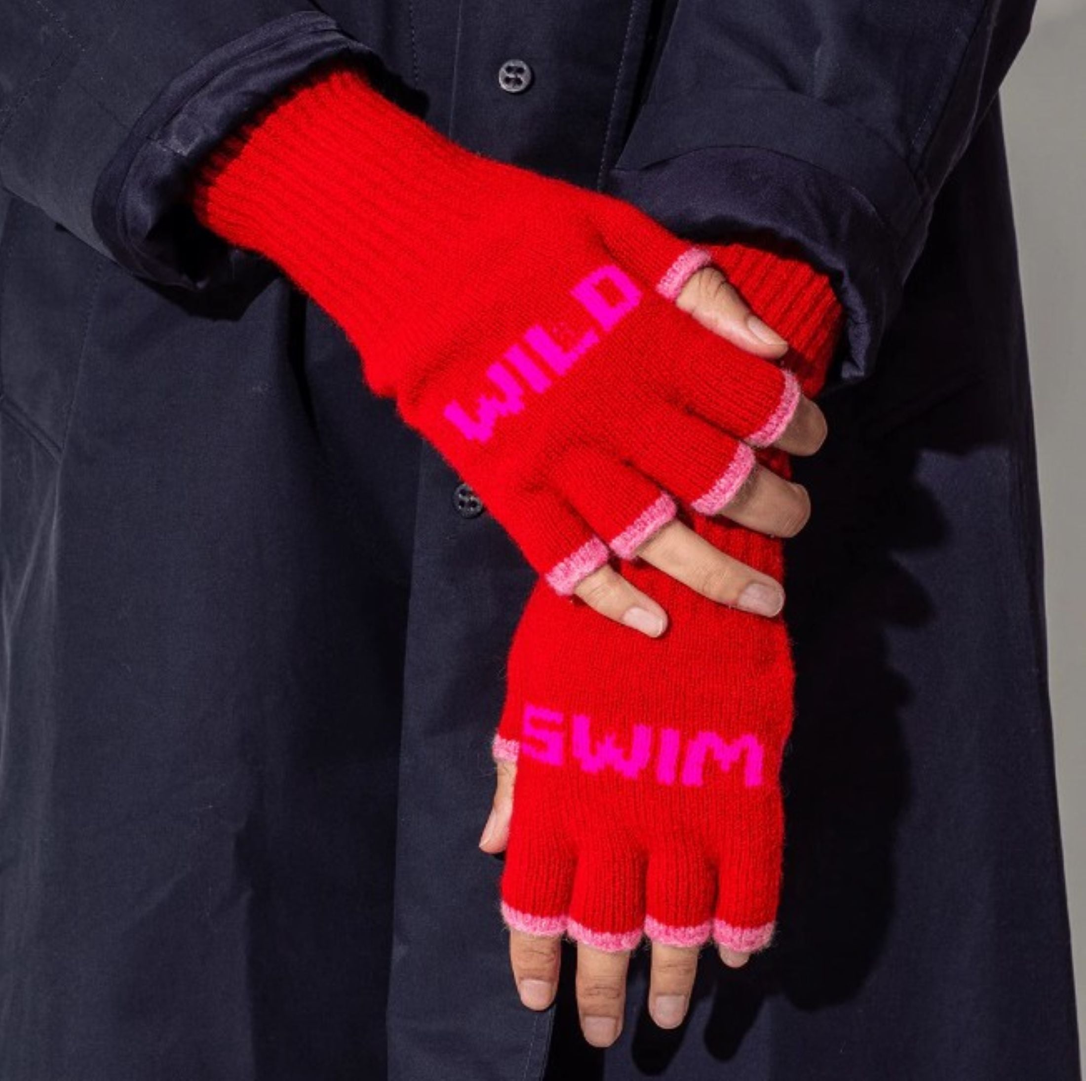 A close up photo of a model wearing the Black & Beech Wild Swim red fingerless gloves with neon pink writing. The model is also wearing a dark blue trench coat.
