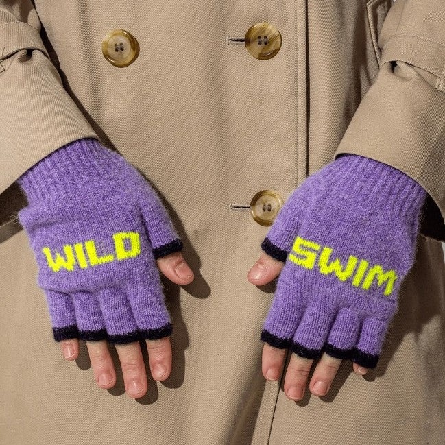 A close up photo of a model wearing the Black & Beech Wild Swim gloves in purple with neon yellow writing and dark purple cuffs. The model is wearing a beige trench coat with tortoise shell buttons.