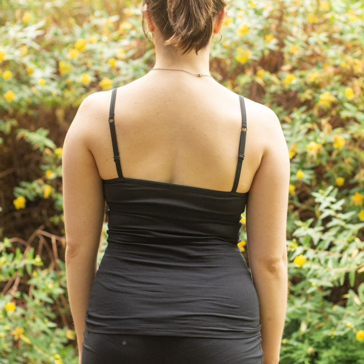 Product focused image of a model wearing the YOU Underwear Organic Cotton Black Camisole. The model faces away from the camera, with her arms by her side. She stands in front of a bush with yellow flowers. The image is cropped from above the ears to the hips.