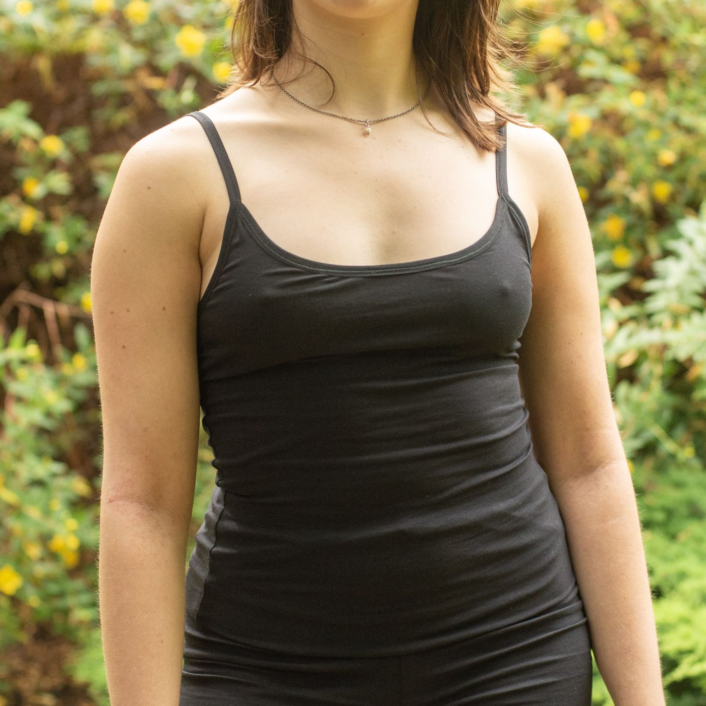 Product focused image of a model wearing the YOU Underwear Organic Cotton Black Camisole. The model faces the camera, with both arms by her side. There is greenery in the background. The image is cropped from the chin to the hips.