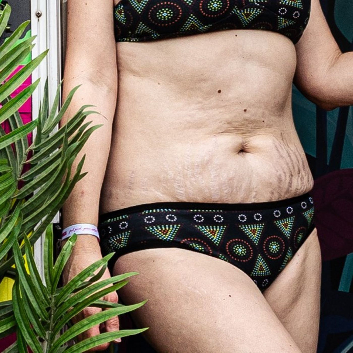 A photo of a model wearing the YOU Underwear low rise bikini knickers in black mara. The photo is zoomed in so we can see from the model's chest to their mid-thigh. They appear to be leaning against a doorway and there is a green plant in the foreground.