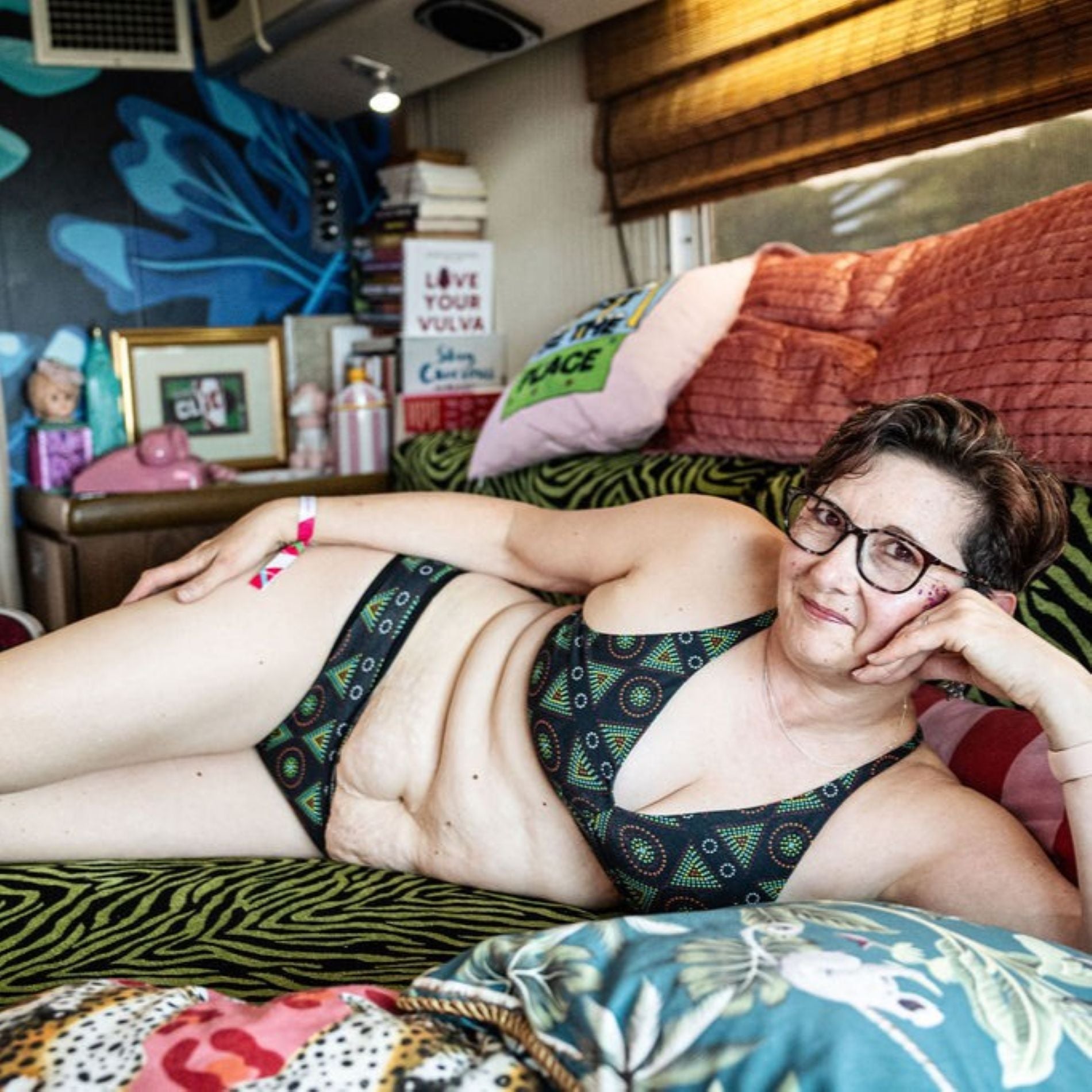 A photo of a model wearing the YOU Underwear bralette and low-rise bikini knickers in black mara. She is laying on her side, on a black and green tiger print sofa. She is facing the camera and smiling. Around her there are several colourful cushions and a side table full of books and pictures. 