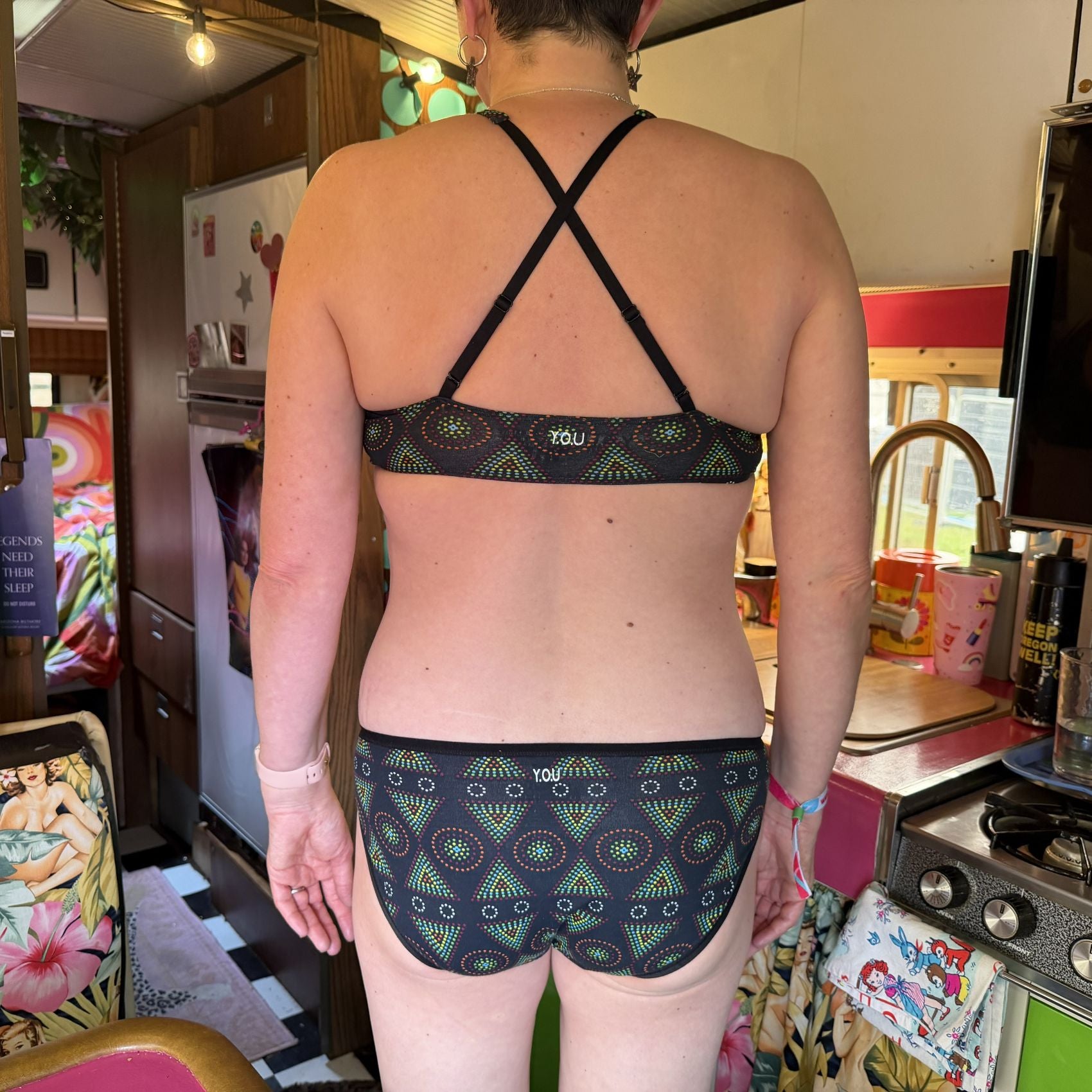 A picture of a model wearing the YOU Underwear Bralette and Low Rise Bikini Knickers in a Black Mara print. They are facing away from the camera with their hands down at their sides. The adjustable straps at the back are crossed to show another style of wear. They are standing in a small colourful kitchen.
