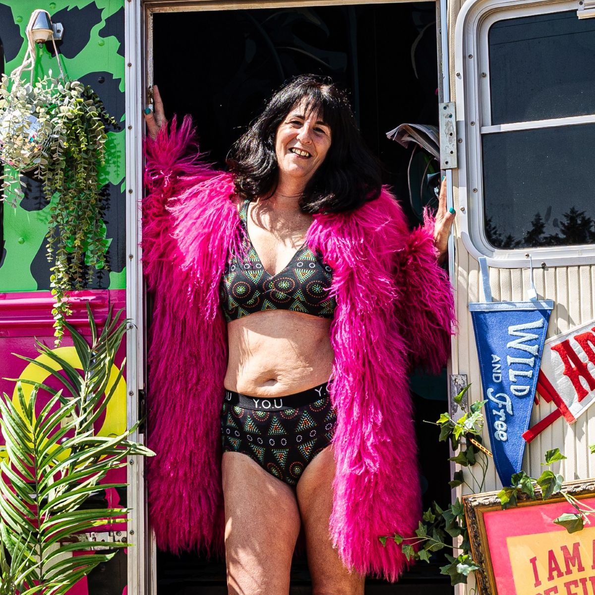 A photo of a model wearing the YOU Underwear bralette and branded boyshorts in the black mara print. She is standing in the doorway of a colourful green and pink bus, with her hands on the door frame. She is wearing a bright pink fluffy coat and standing facing the camera, smiling.